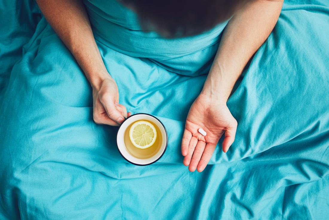 Top down view of person sitting under blue blanket holding mug of lemon tea and pill or supplement