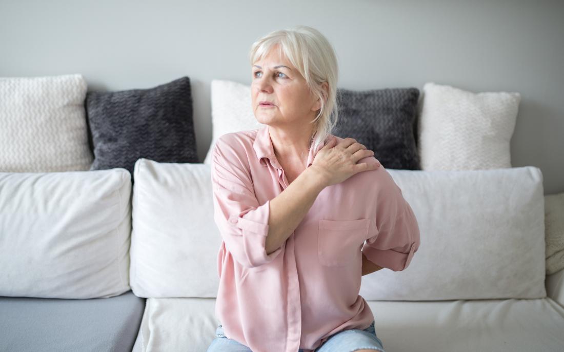 senior woman sitting on settee holding her shoulder in pain