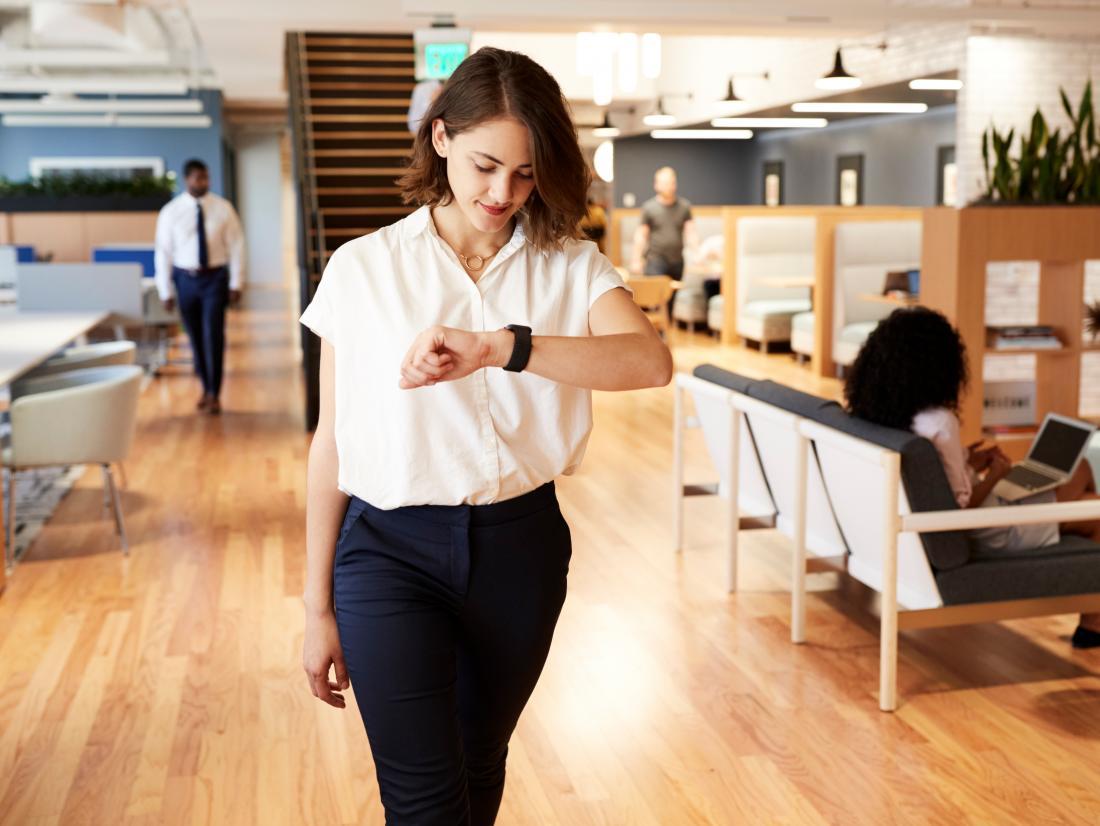 a woman walking around an office and looking at her watch.